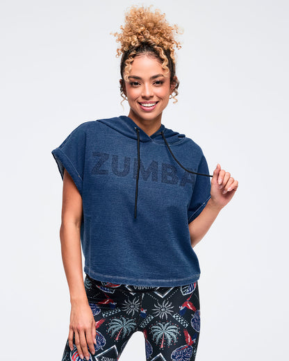 Navy denim cropped short-sleeve hoodie with tonal “ZUMBA” chest graphic and black drawstrings; female model pairs it with dark tropical-print leggings on a white background.