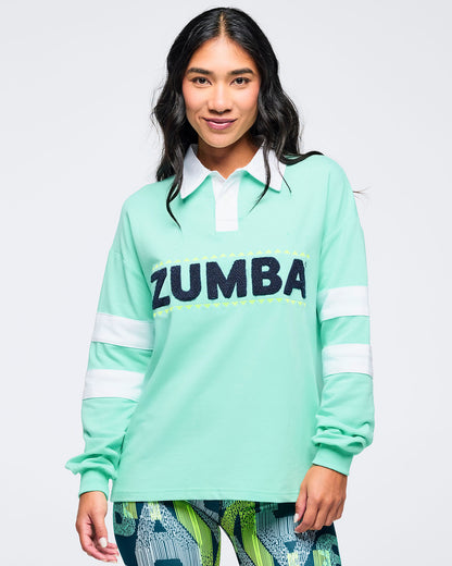 Front view of the mint green rugby sweatshirt with white collar and bold “ZUMBA” chest lettering on a female model, styled with green abstract print bottoms on a white background.