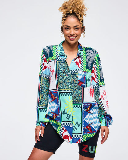 Multicolor patchwork button-up Zumba shirt with tropical panels, checkerboard accents, and parrot prints in teal, navy, red, and lime. Female model wears the long-sleeve collared shirt over black biker shorts with a subtle Zumba logo on a light gray studio background.