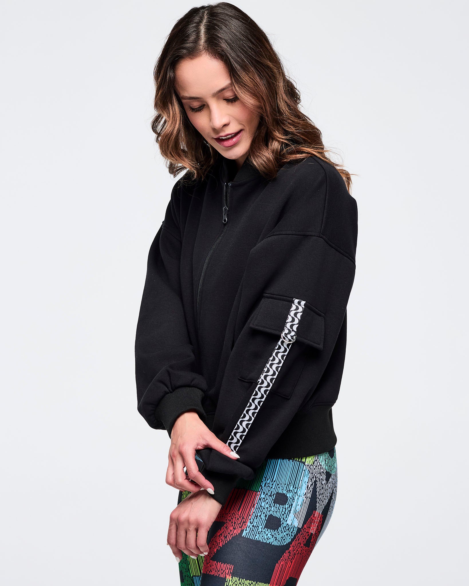 Black zip-up jacket featuring a sleeve cargo pocket and black-and-white wavy trim detail; female model wears multicolor letter-print leggings on a white background.