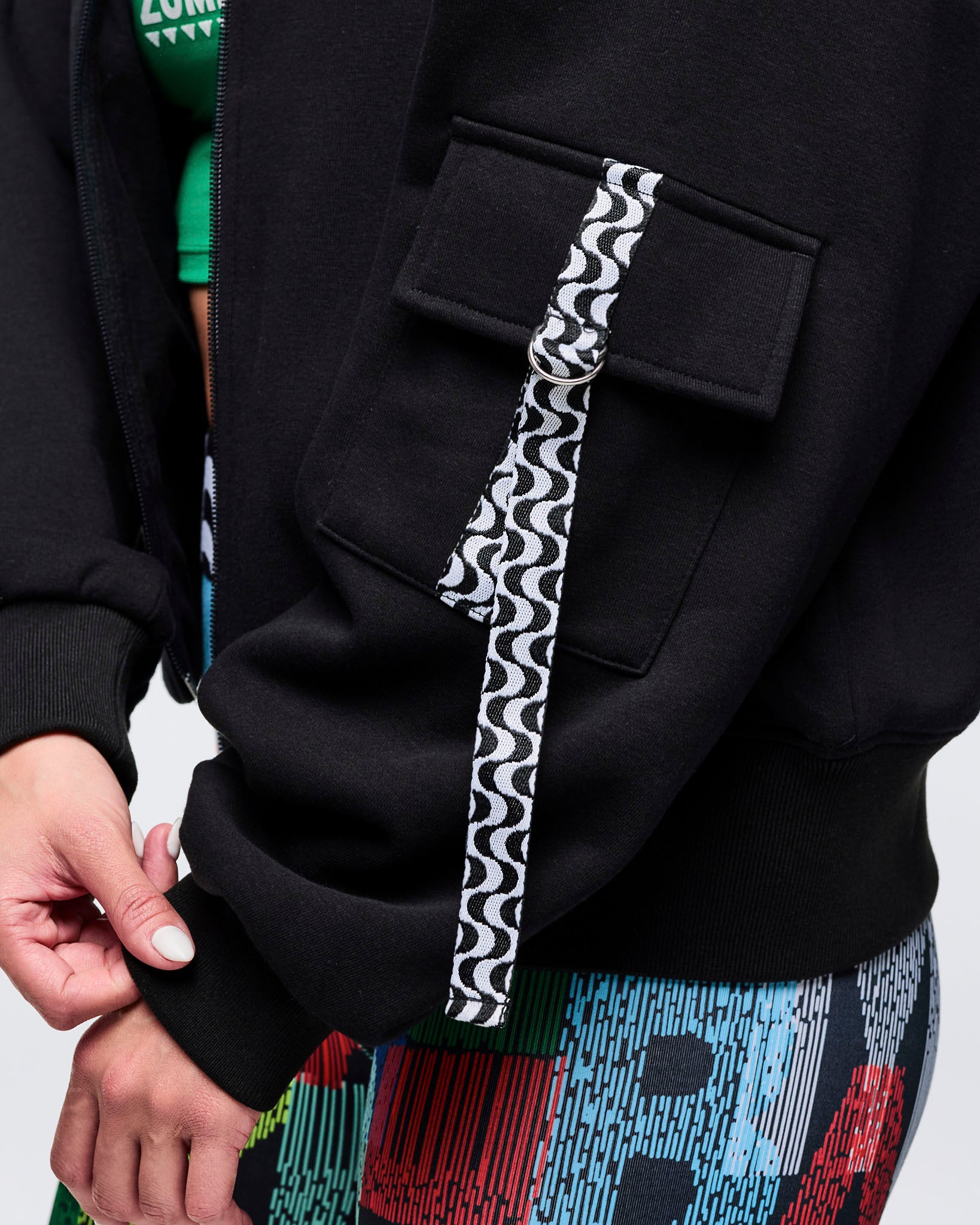 Close-up of the zip-up jacket sleeve showing the flap pocket and black-and-white wavy trim accent beside the zipper; female model wears multicolor letter-print leggings.