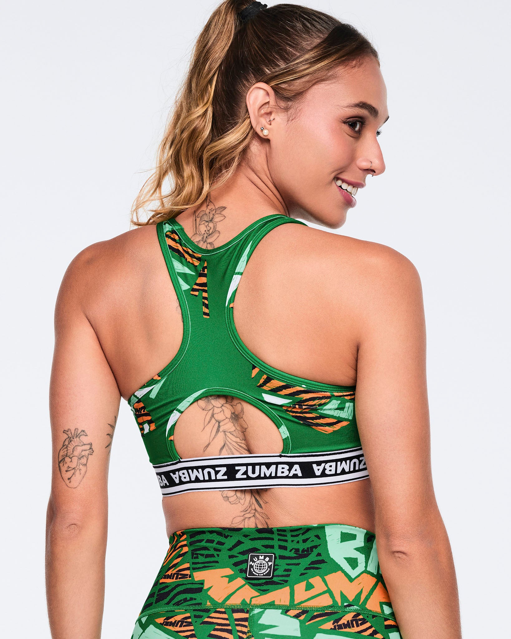 Female model wearing a green racerback sports bra shown from the back with a keyhole cutout, a black-and-white Zumba logo band, and an abstract print in green, mint, orange, black, and white.