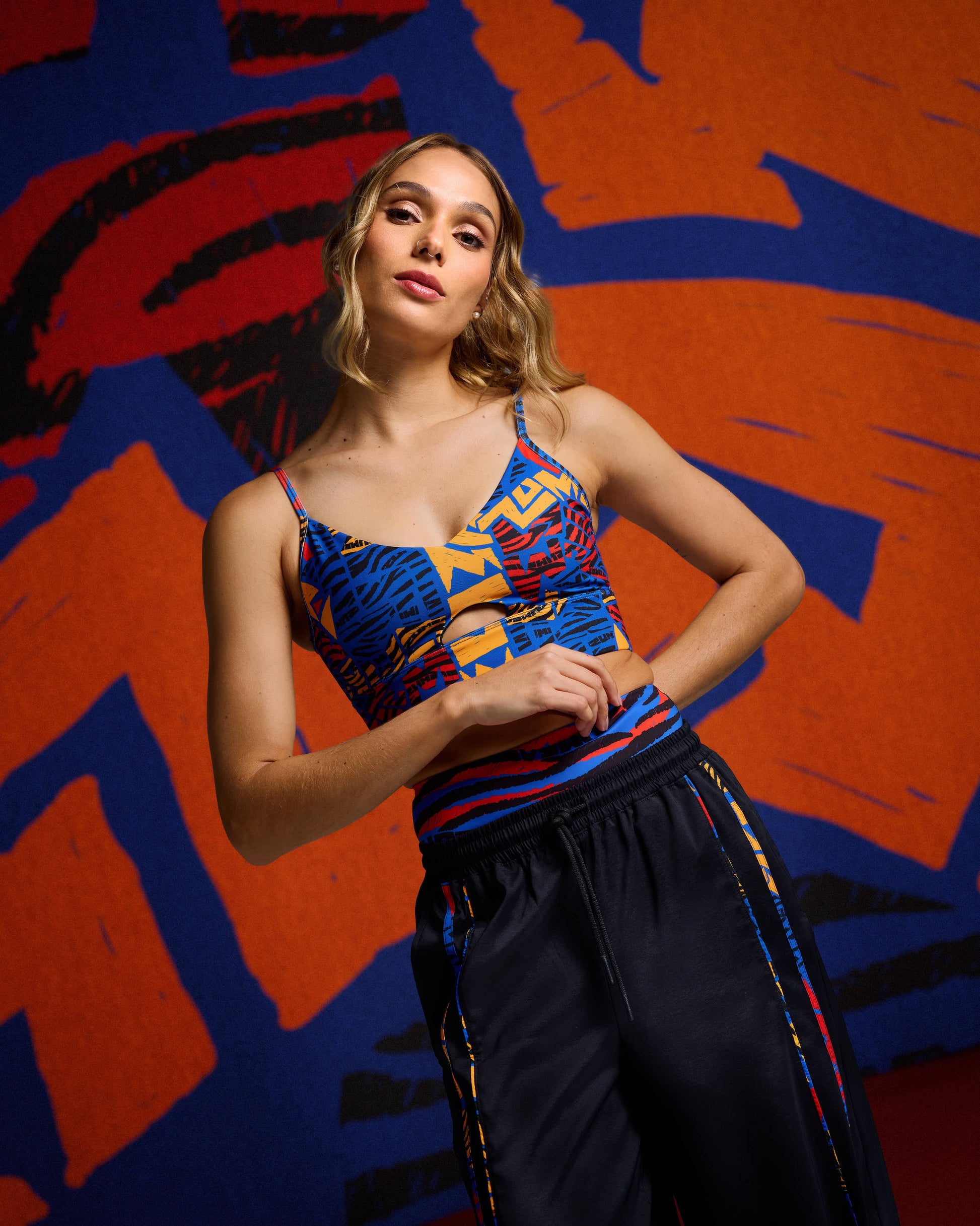 Female model wearing a cropped strappy sports bra in a bold blue, orange, red, and black abstract print, paired with black wide-leg track pants with matching printed trim and an elastic waistband.