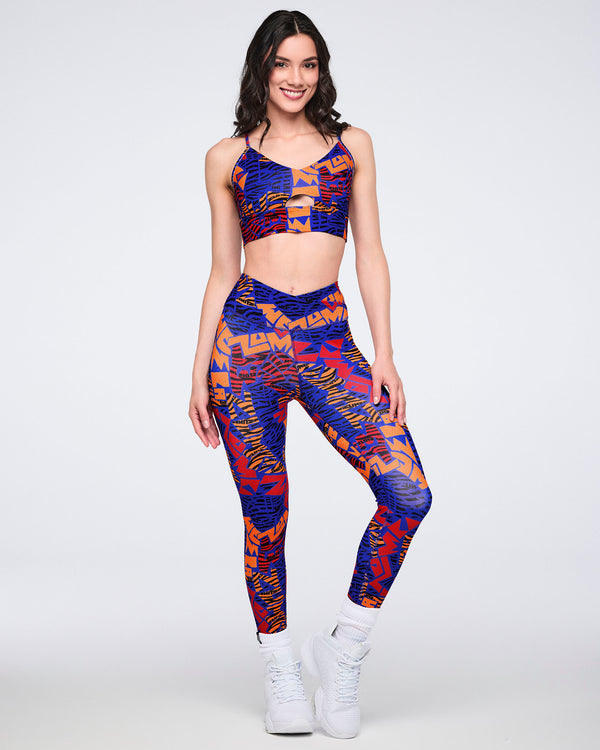 Female model wearing a matching set with a cropped strappy sports bra and high-waisted full-length leggings in a blue, orange, red, and black abstract print, paired with white high-top sneakers.
