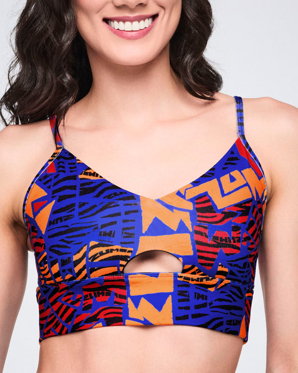 Female model wearing a cropped strappy sports bra in a blue, orange, red, and black abstract print with a front keyhole cutout and a fitted longline silhouette.
