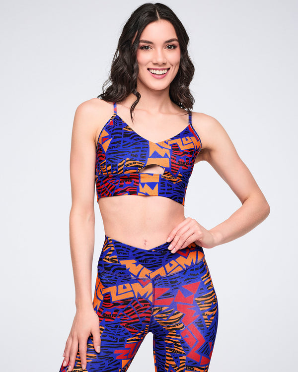 Female model wearing a matching set with a cropped strappy sports bra featuring a front keyhole cutout and high-waisted leggings in a blue, orange, red, and black abstract print.