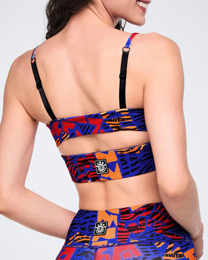 Female model wearing a cropped strappy sports bra in a blue, orange, red, and black abstract print, shown from the back with adjustable black shoulder straps and a wide printed back band.