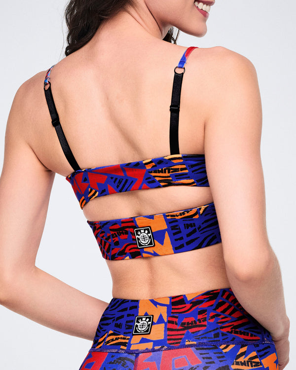 Female model wearing a cropped strappy sports bra in a blue, orange, red, and black abstract print, shown from the back with adjustable black shoulder straps and a wide printed back band.