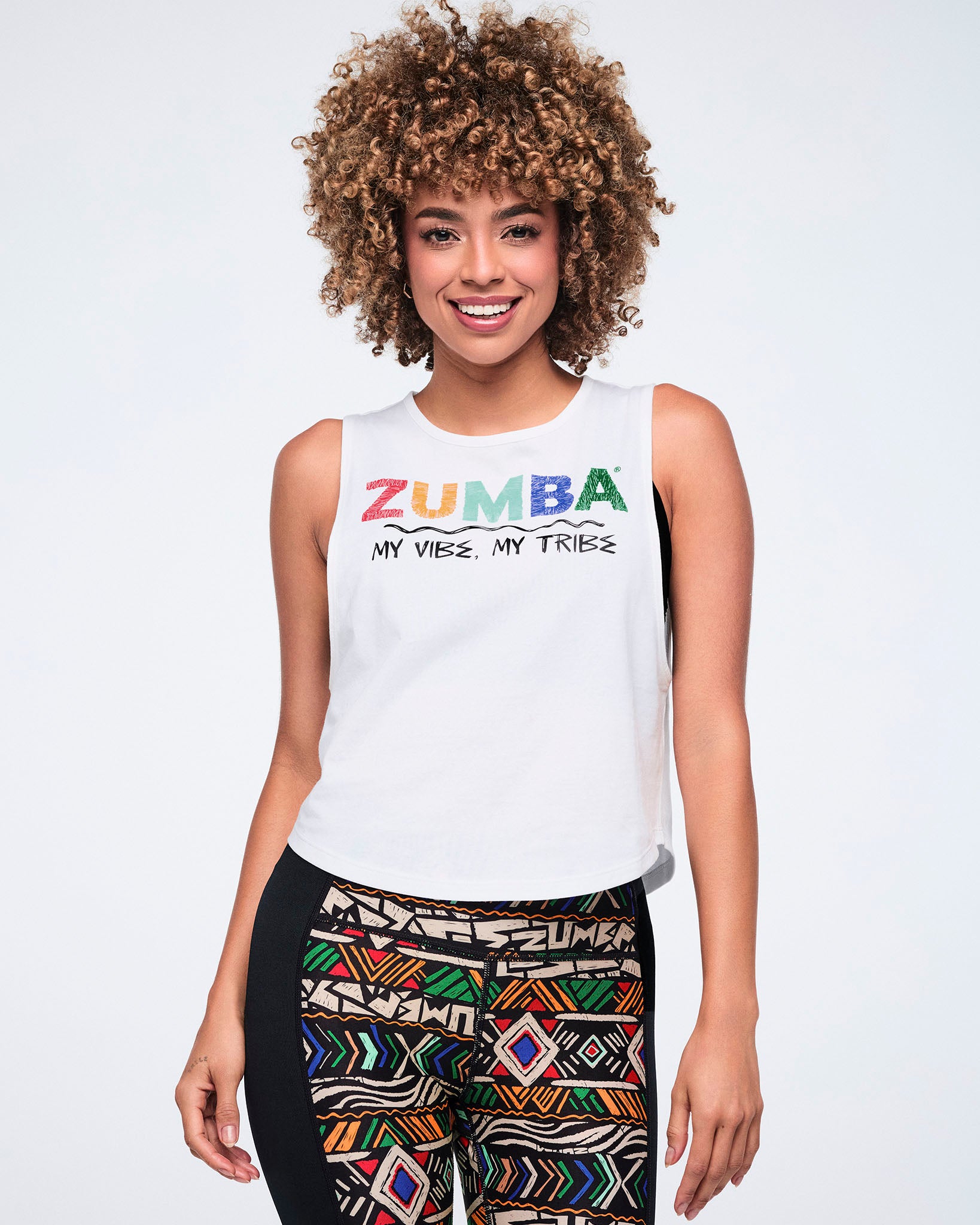 Female model wearing a white sleeveless tank with multicolor Zumba text and “My Vibe, My Tribe” printed on the front, paired with black leggings featuring a bold multicolor geometric print and black side panels.