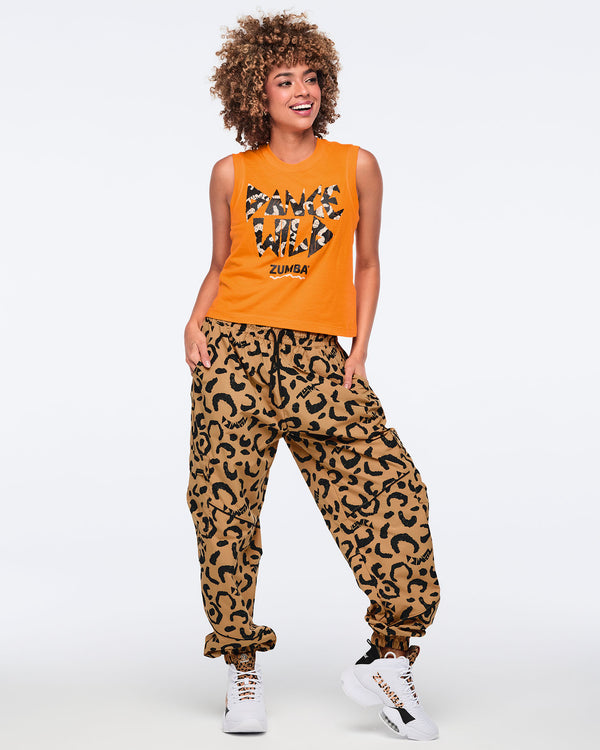 Orange sleeveless tank top with bold “Dance Wild” graphic lettering and a black Zumba logo, shown on a smiling female model wearing tan leopard-print cargo pants and white high-top sneakers with leopard-print details.