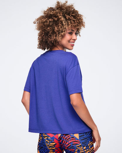 Back view of the blue Zumba graphic T-shirt with a relaxed fit, crew neckline, and short sleeves. Female model wears the tee with multicolor abstract-print leggings, shown turned over her shoulder.