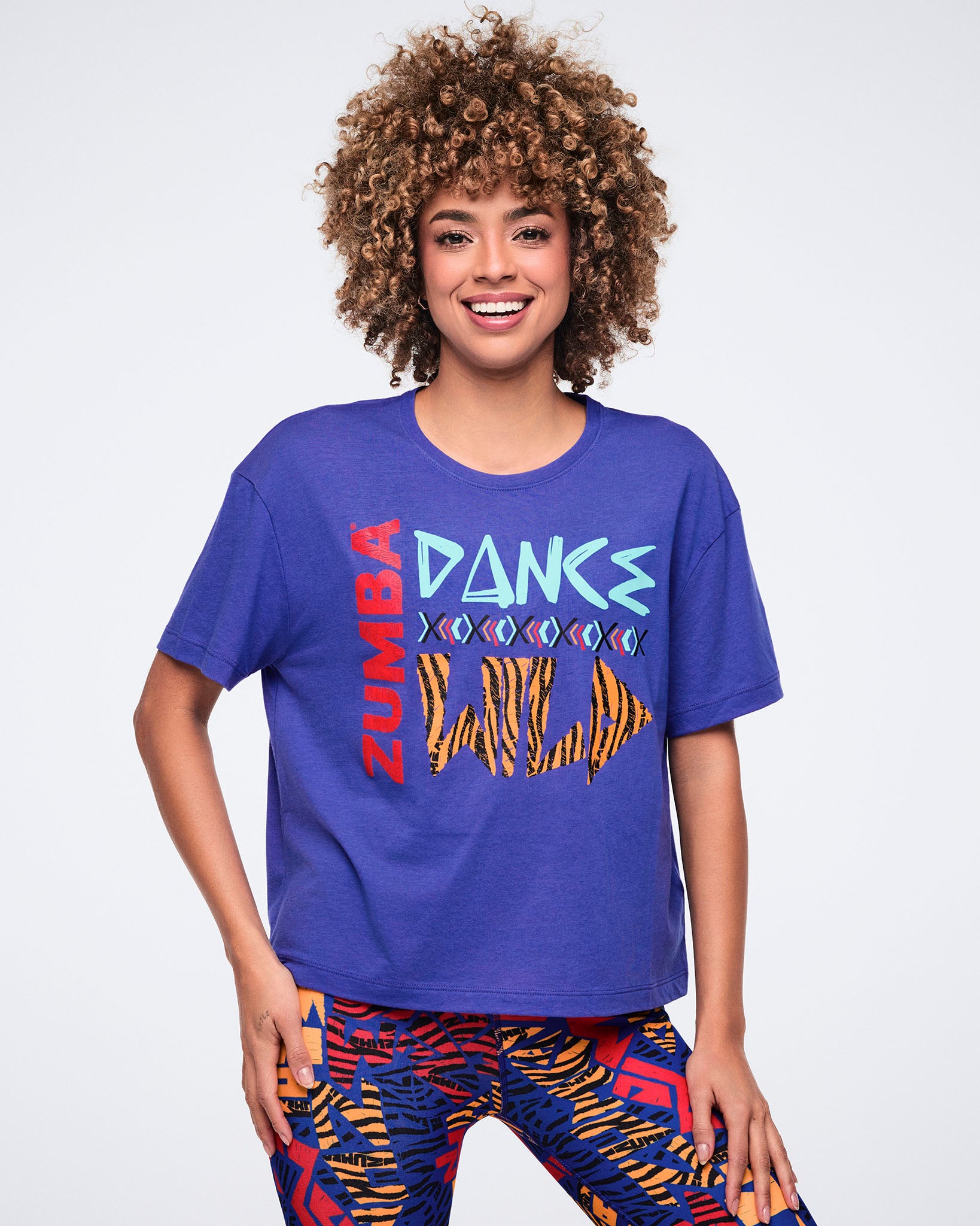 Blue Zumba graphic T-shirt with a relaxed fit, crew neckline, and short sleeves, featuring a bold front print with red “ZUMBA,” light blue “DANCE,” and an orange-and-black “WILD” graphic. Female model wears the tee with multicolor abstract-print leggings.
