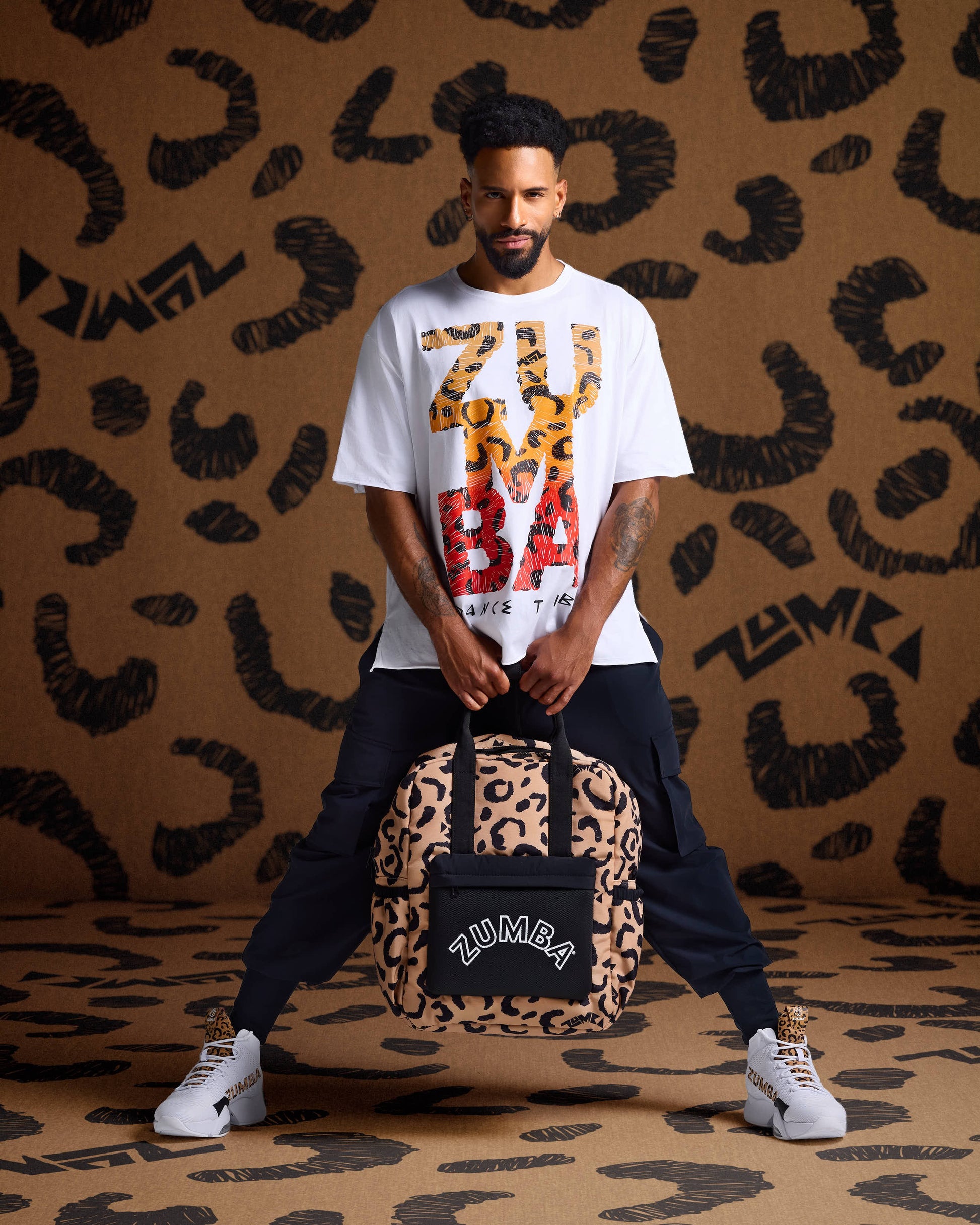 Leopard print backpack held by a male model in a white graphic tee, black cargo joggers, and white high-top sneakers against  leopard print backdrop.