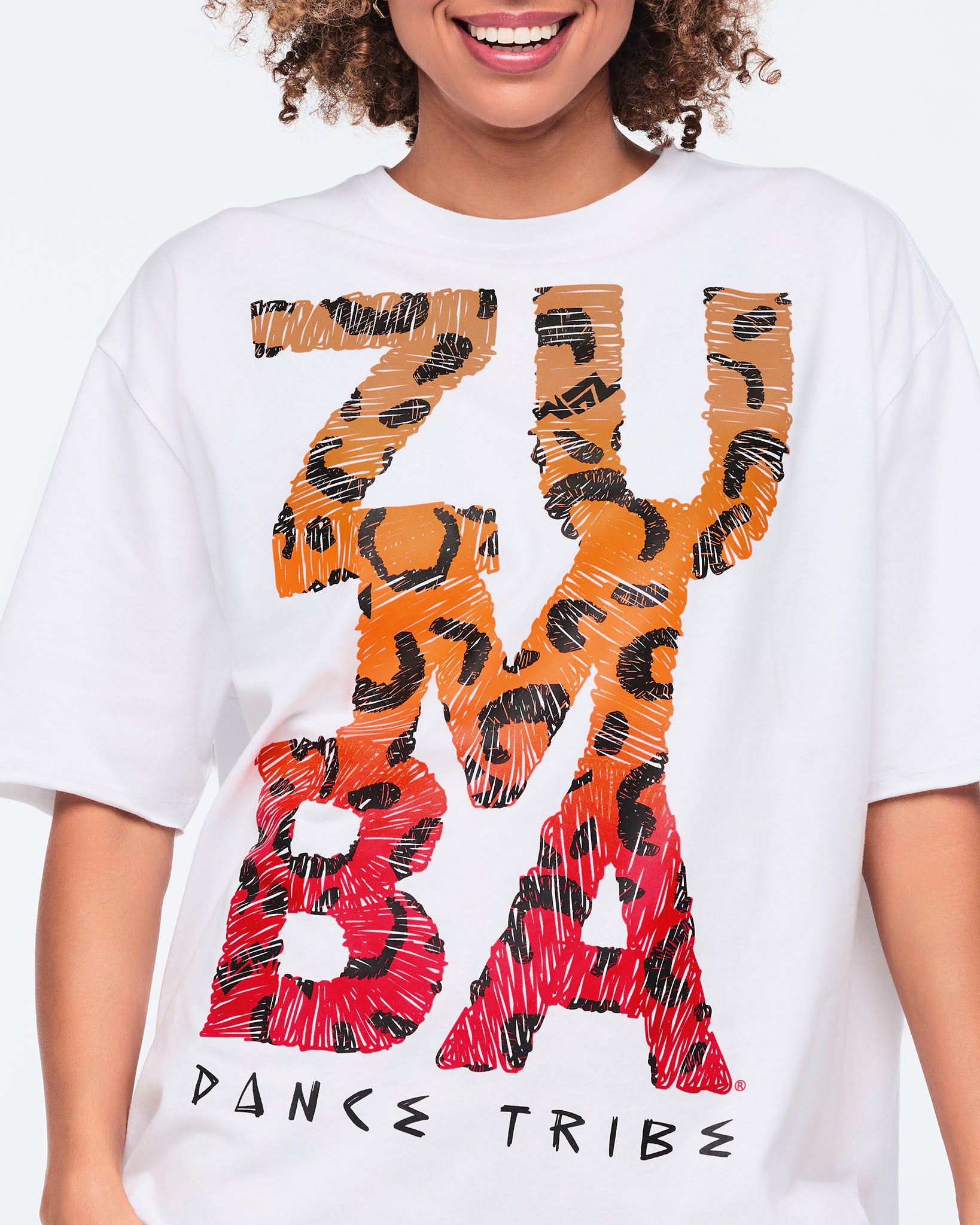 White oversized T-shirt with large orange and red leopard-print “Zumba Dance Tribe” graphic lettering, shown in close-up on a female model.