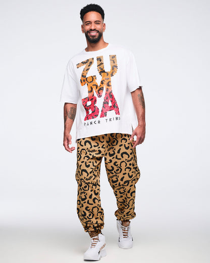White oversized T-shirt with large multicolor “Zumba Dance Tribe” graphic lettering, shown on a smiling male model wearing tan leopard-print cargo pants and white high-top sneakers with leopard-print details.