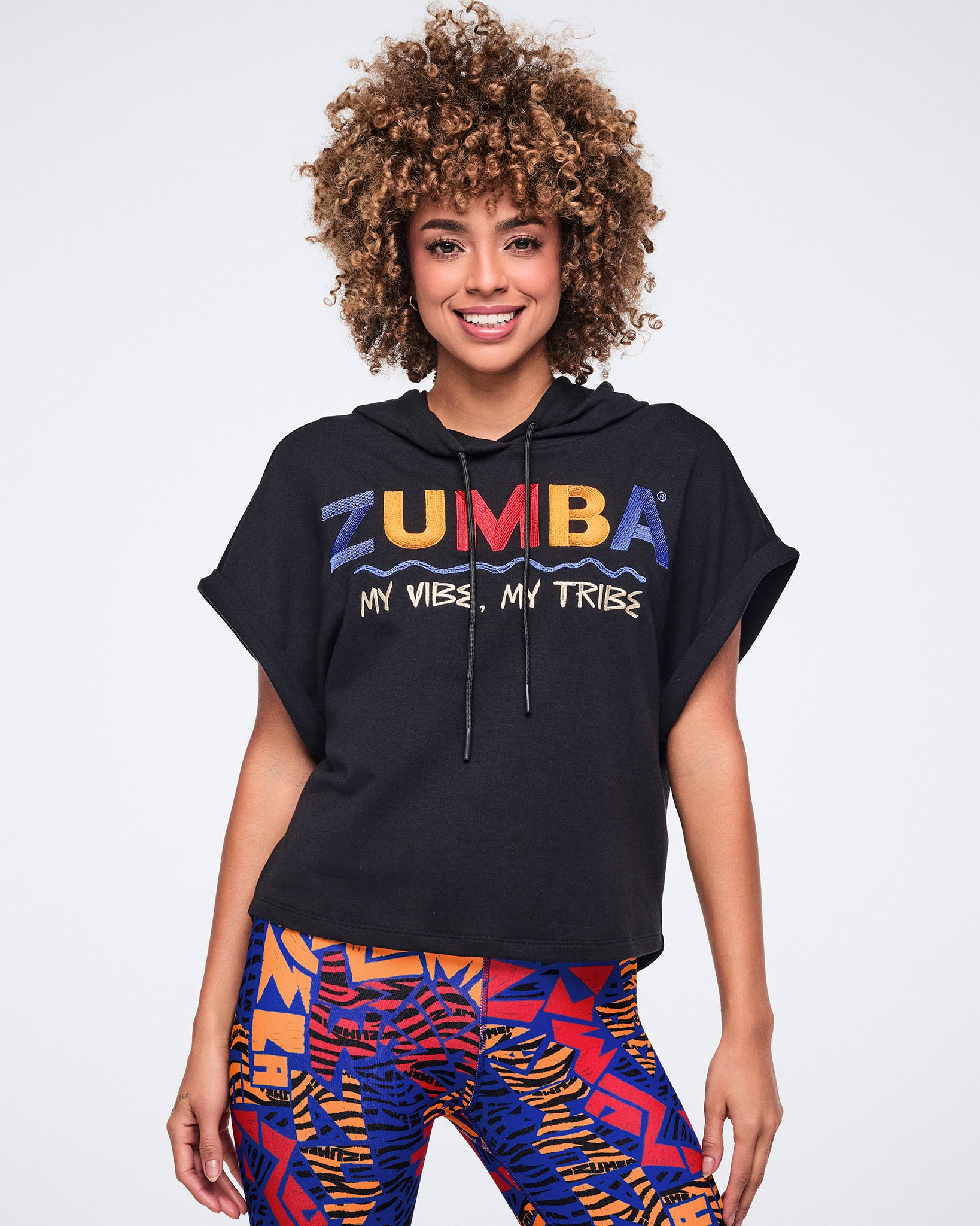 Black short-sleeve hoodie with rolled cuffs, a drawstring hood, and multicolor embroidered Zumba graphic text reading “My Vibe, My Tribe” across the chest. Female model wears the hoodie with bright abstract-print leggings, standing and smiling against a light gray background.