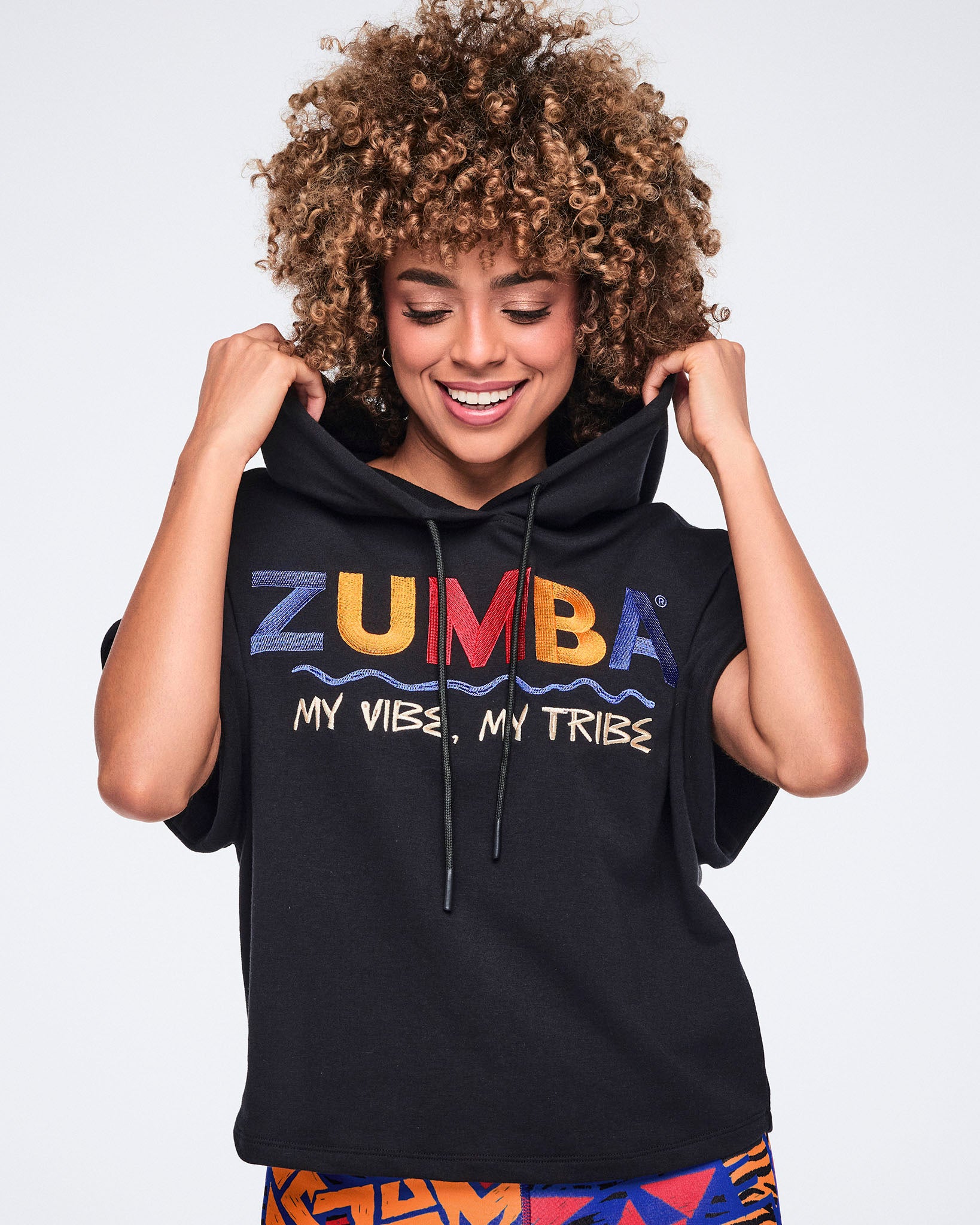 Female model wears the hoodie with bright abstract-print leggings, smiling while holding the sides of the hood against a light gray background .Model wears a black short-sleeve hoodie with rolled cuffs, a drawstring hood, and multicolor embroidered Zumba graphic text reading “My Vibe, My Tribe” across the chest. 