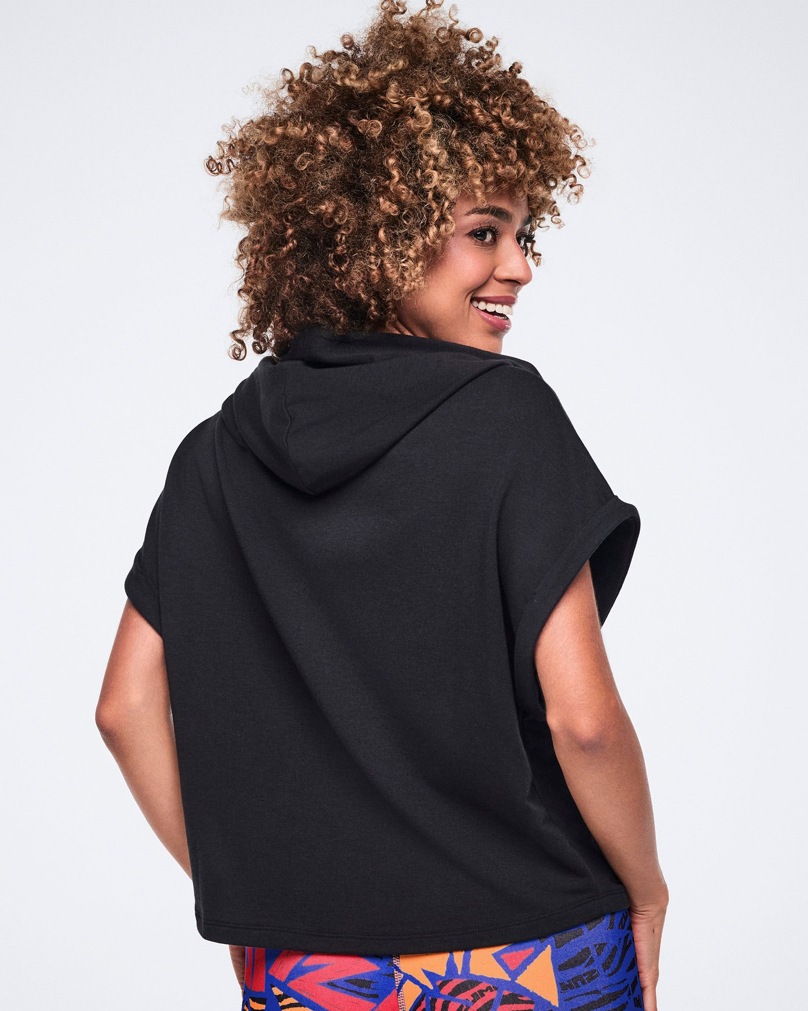 Female model wears the hooded top with bright abstract-print leggings, shown from the back while looking over her shoulder against a light gray background.