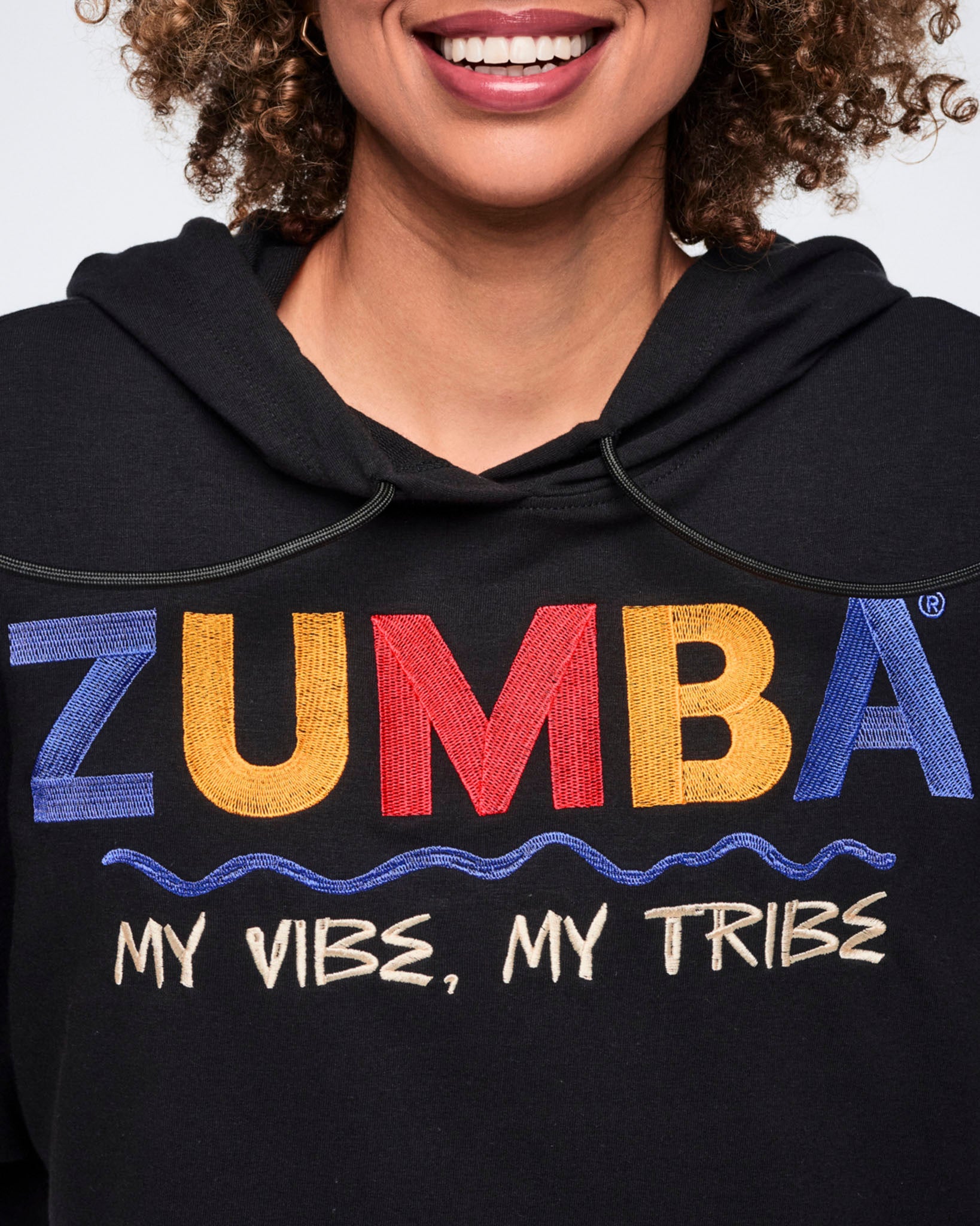 Close-up of a black short-sleeve hoodie with a drawstring hood and multicolor embroidered Zumba graphic text reading “My Vibe, My Tribe” across the chest. The image highlights the blue, yellow, and red Zumba lettering and cream slogan detail on the front.