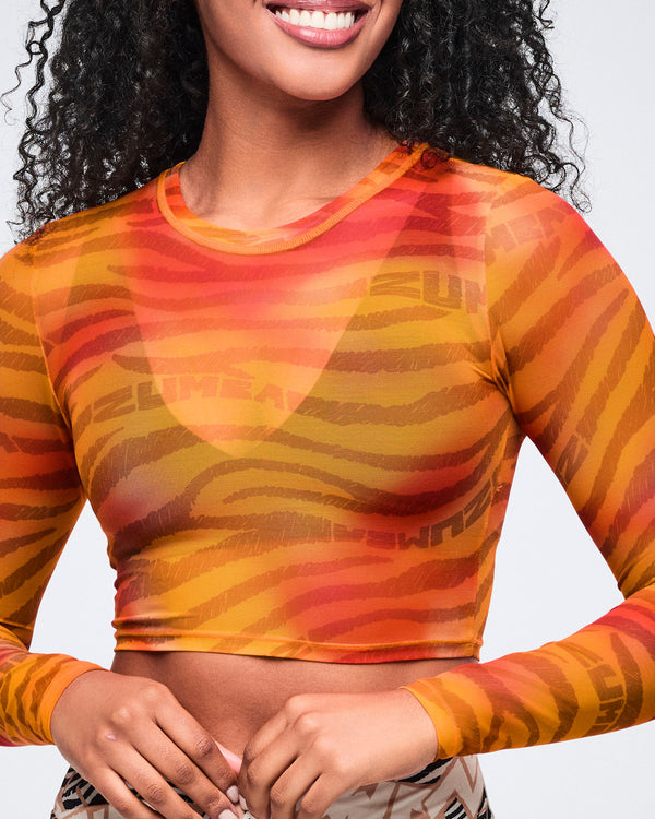 Close-up of an orange and red zebra-print sheer long-sleeve crop top with a crew neckline and fitted shape. Female model wears the top over a matching orange bralette with a beige, black, and cream zebra-print skirt.