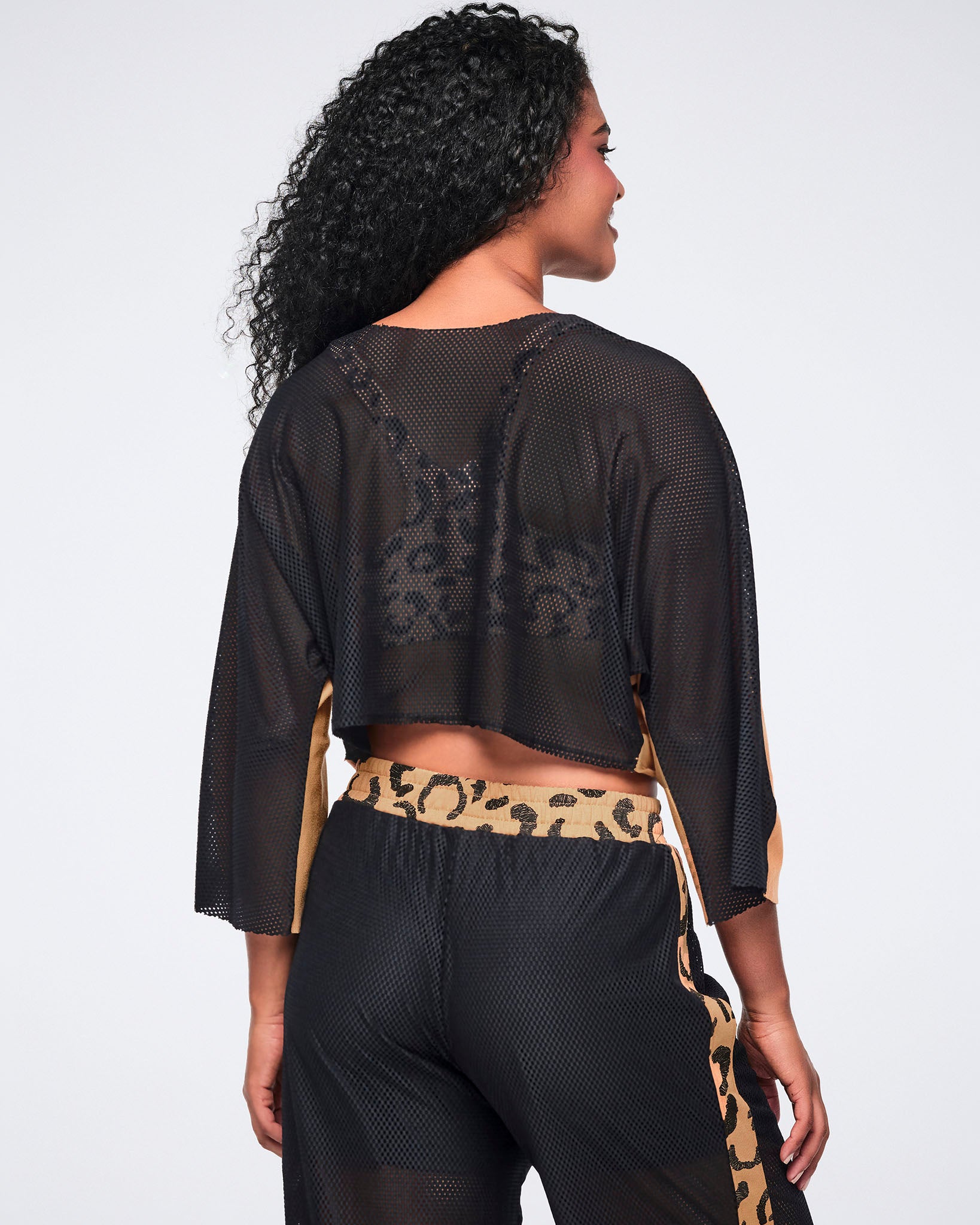 Back of a french terry knit top showing the black mesh back with a sheer finish and loose fit, on a female model wearing black mesh pants with a tan cheetah-print accents.