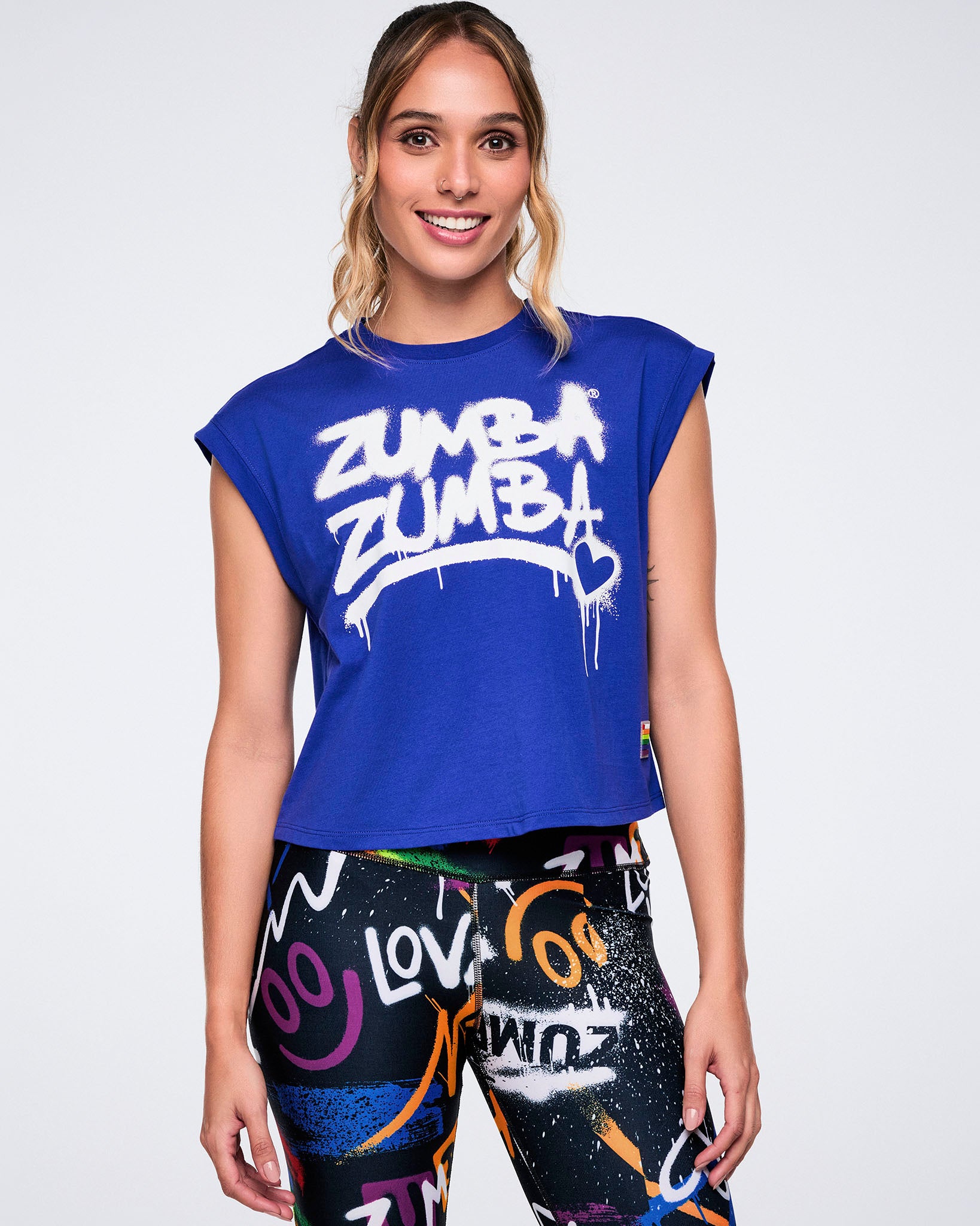 Female model in a blue Zumba top with white spray‑painted “Zumba Zumba” and heart detail, paired with colorful leggings on a white background.