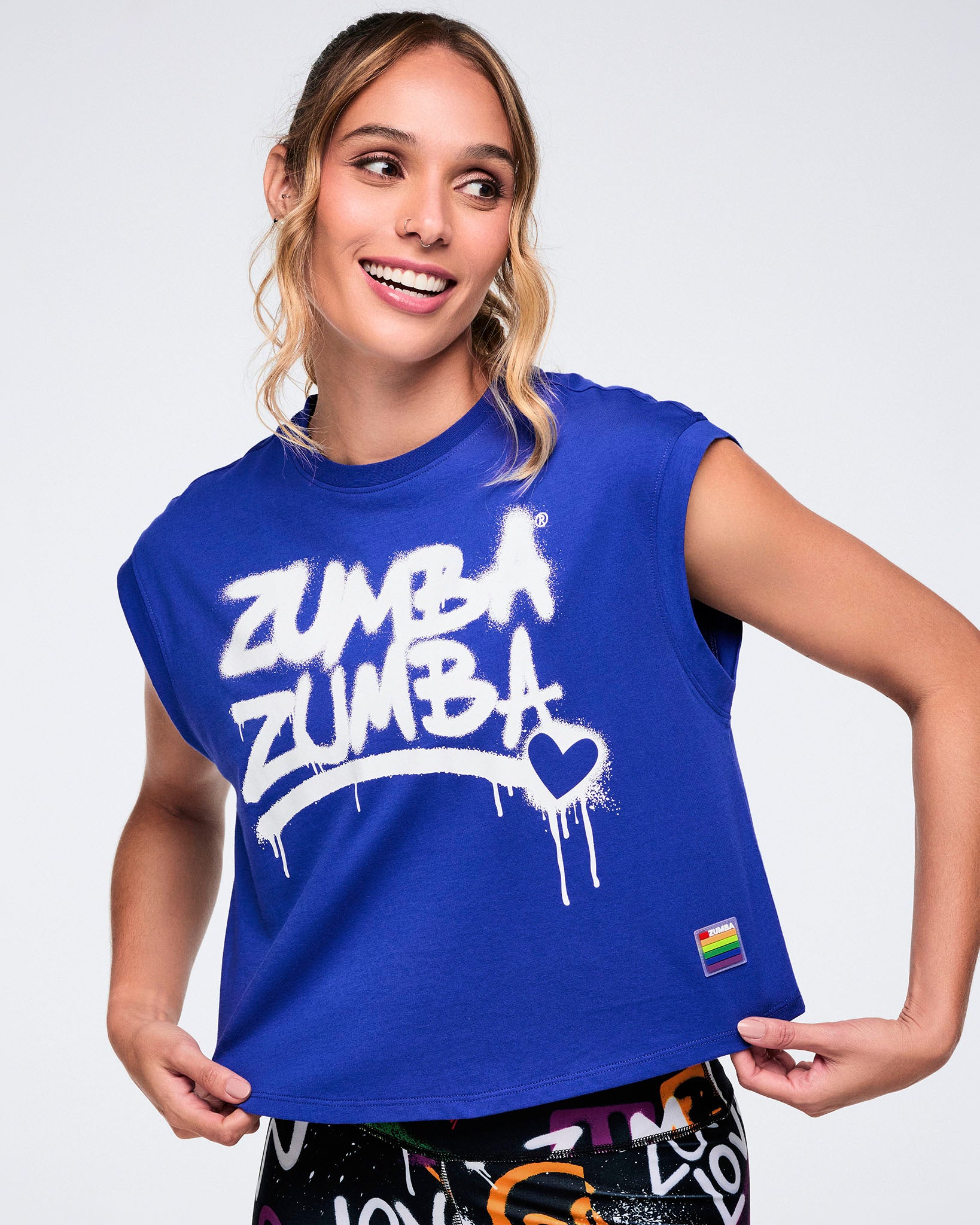 Female model with a close up showing off a blue Zumba top with white spray‑painted “Zumba Zumba” with heart detail, and a badge on the lower left of the shirt, paired with colorful leggings on a white background.