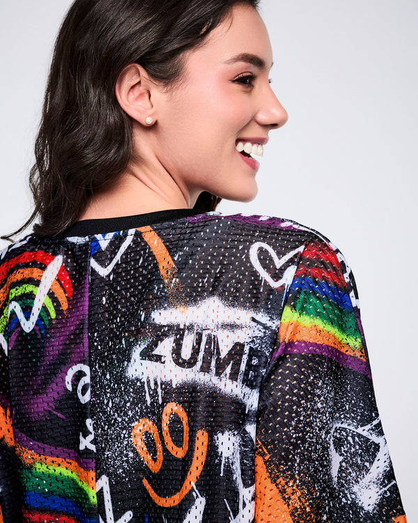 Close up of a female model wearing a colorful, mesh crop top with graffiti-style designs on a plain background.