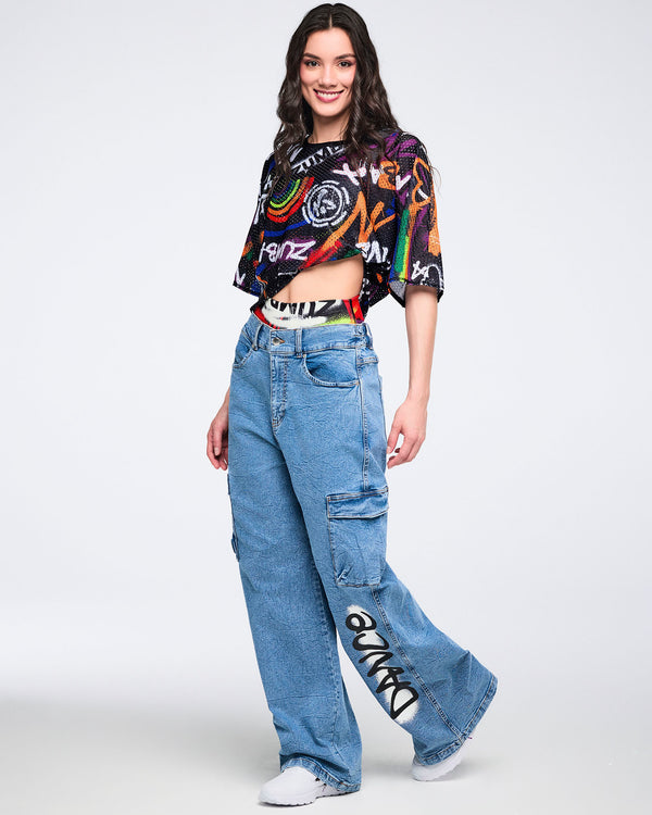 Full body view of a female model wearing a colorful mesh crop top and denim cargo pants on a white background.