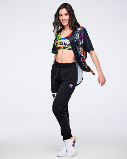 Female model in colorful Zumba crop top, black joggers, and white sneakers posing against a white background.