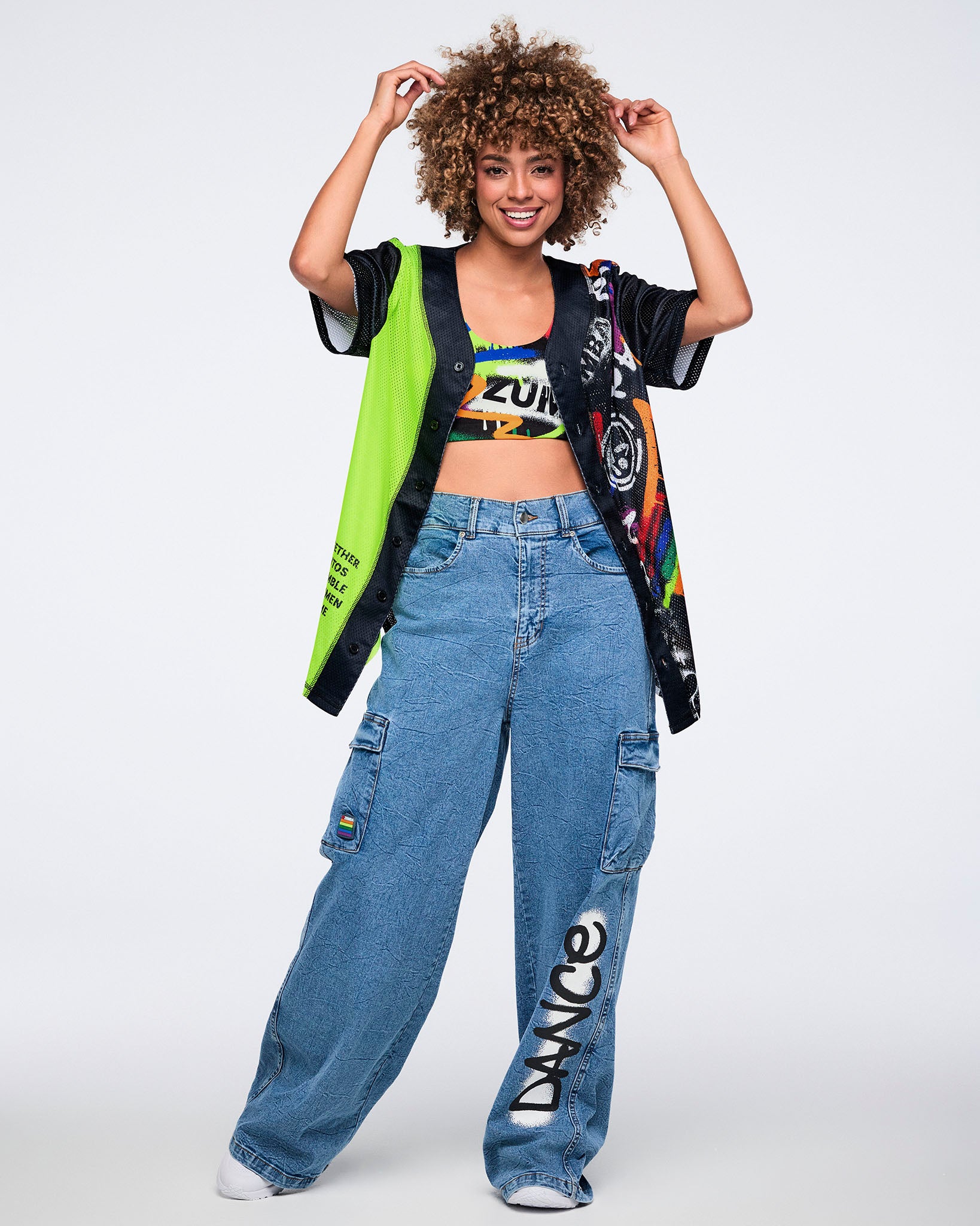 Female model in colorful Zumba bra, a neon‑green and black jersey, and denim cargo pants with the word 'DANCE' printed vertically on the left leg, all against a white background.