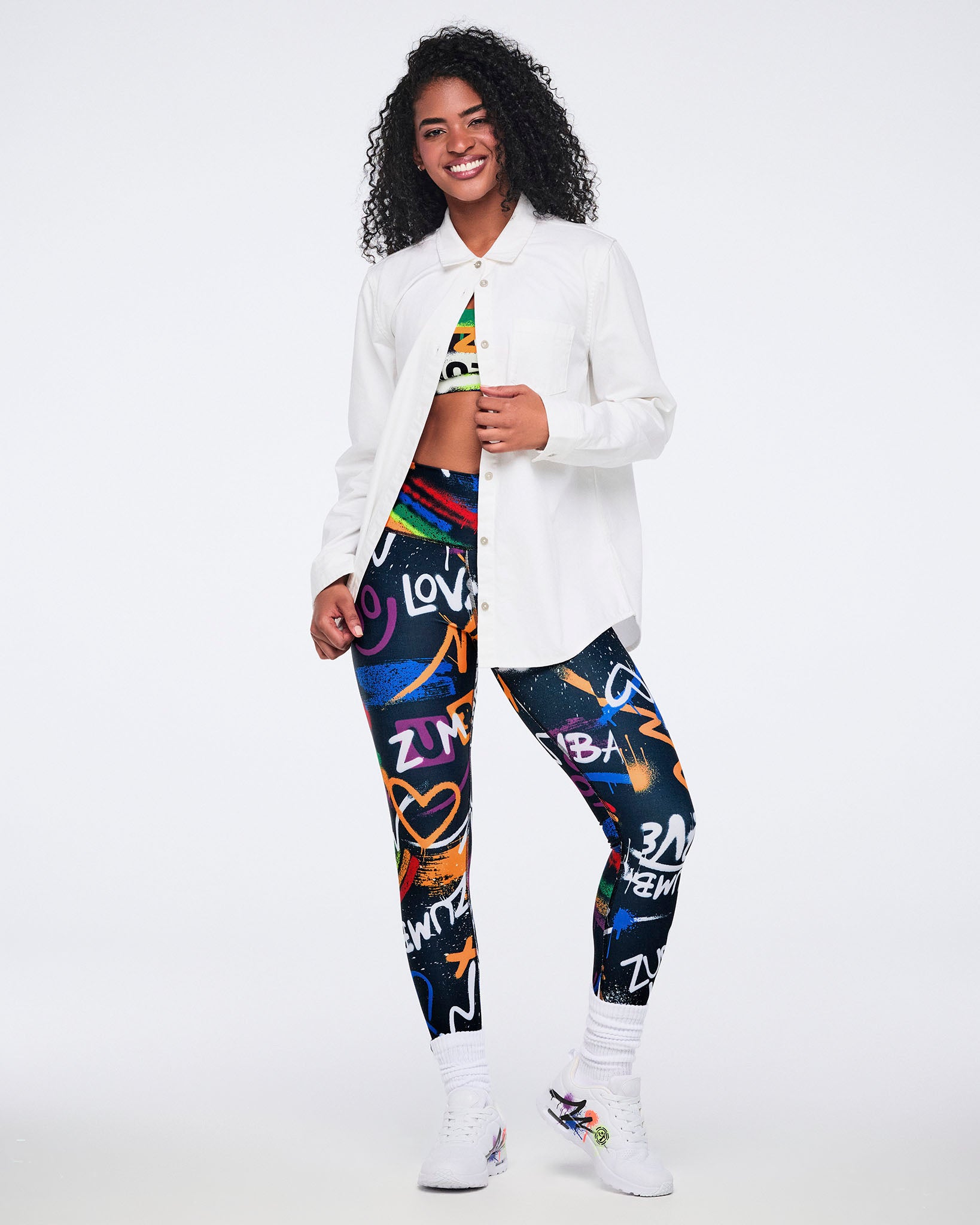 Female model wearing a white button‑down shirt fastened at the top, revealing a colorful matching bra and leggings, paired with white graffiti‑style Zumba sneakers against a white background.