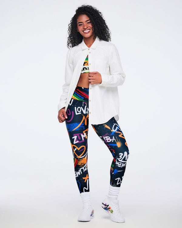 Female model wearing a white button‑down shirt fastened at the top, revealing a colorful matching bra and leggings, paired with white graffiti‑style Zumba sneakers against a white background.