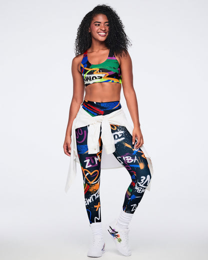 Female model wearing a colorful sports bra and matching leggings, with a white button-down tied around her waist and white Zumba shoes against a white background.
