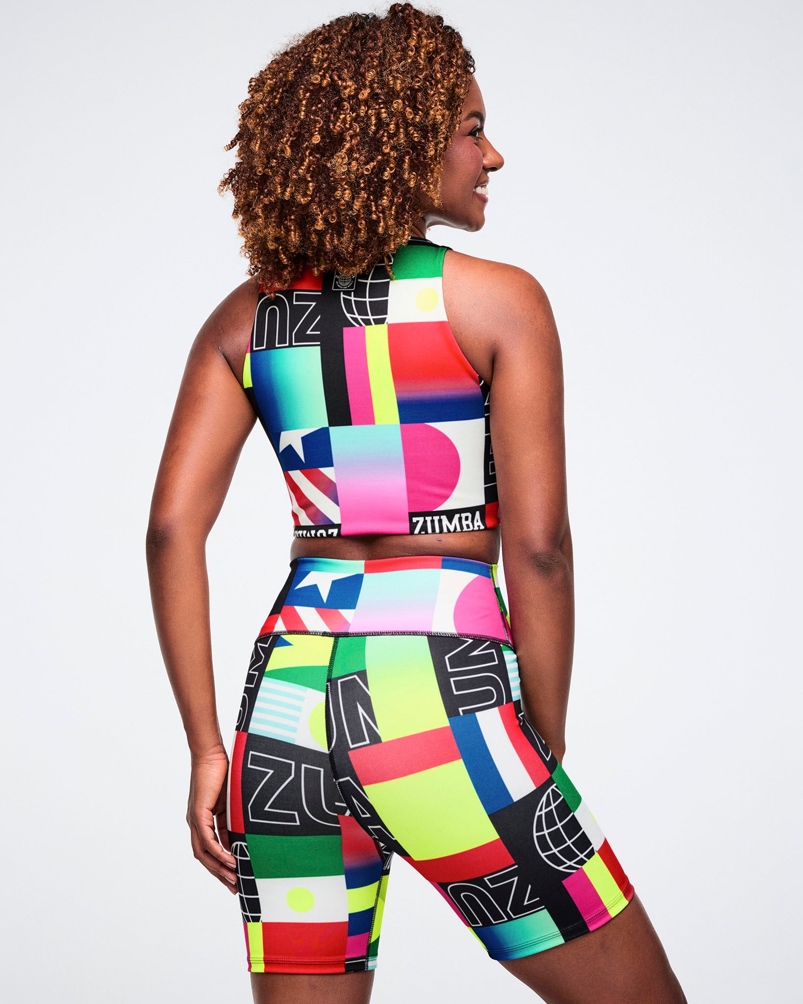 Back view of a female model wearing a colorful crop top and biker shorts with geometric patterns and brand logos on a plain background.