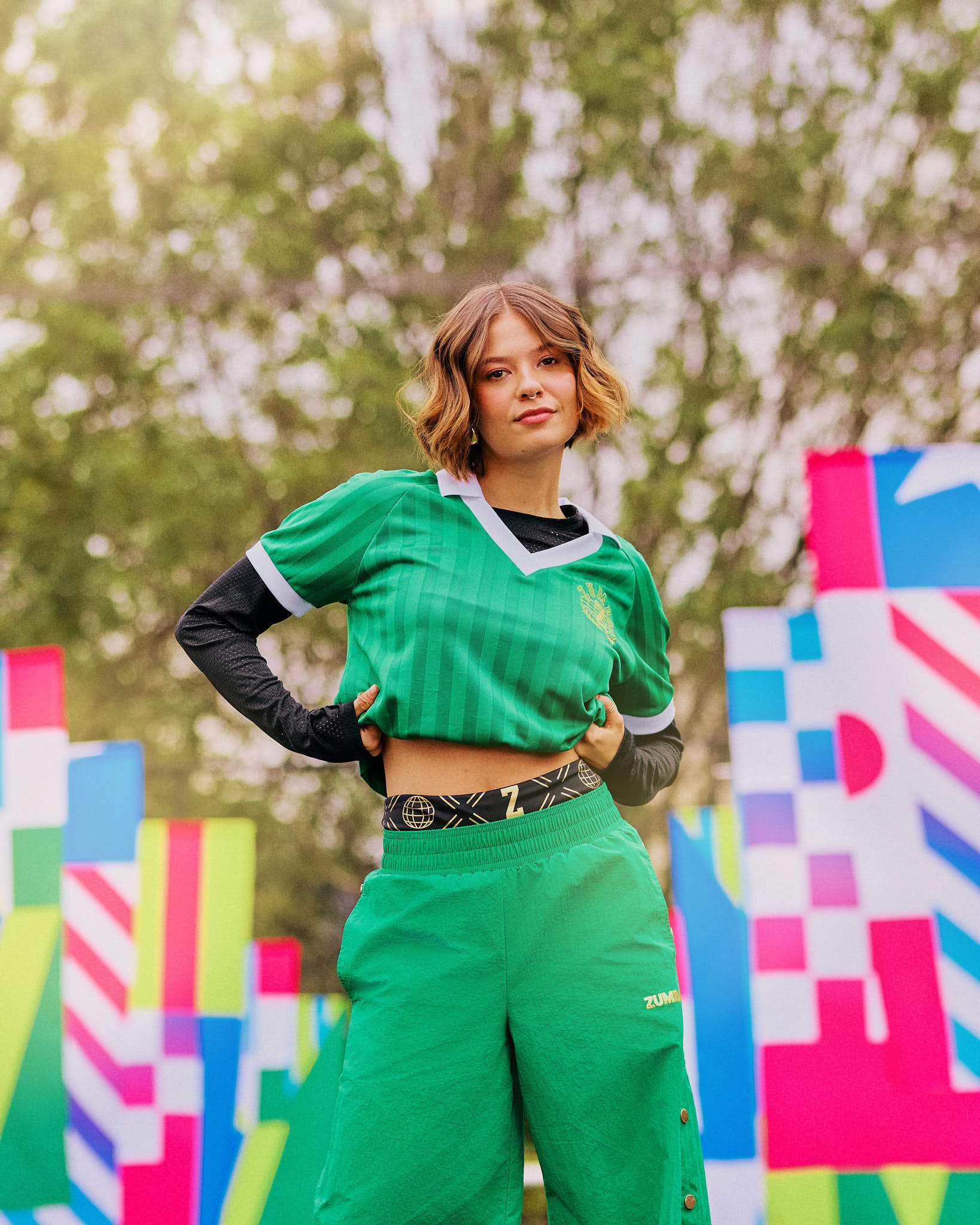 Female model outdoors in a green polo and green snapback pants, with colorful geometric flag patterns in the background.