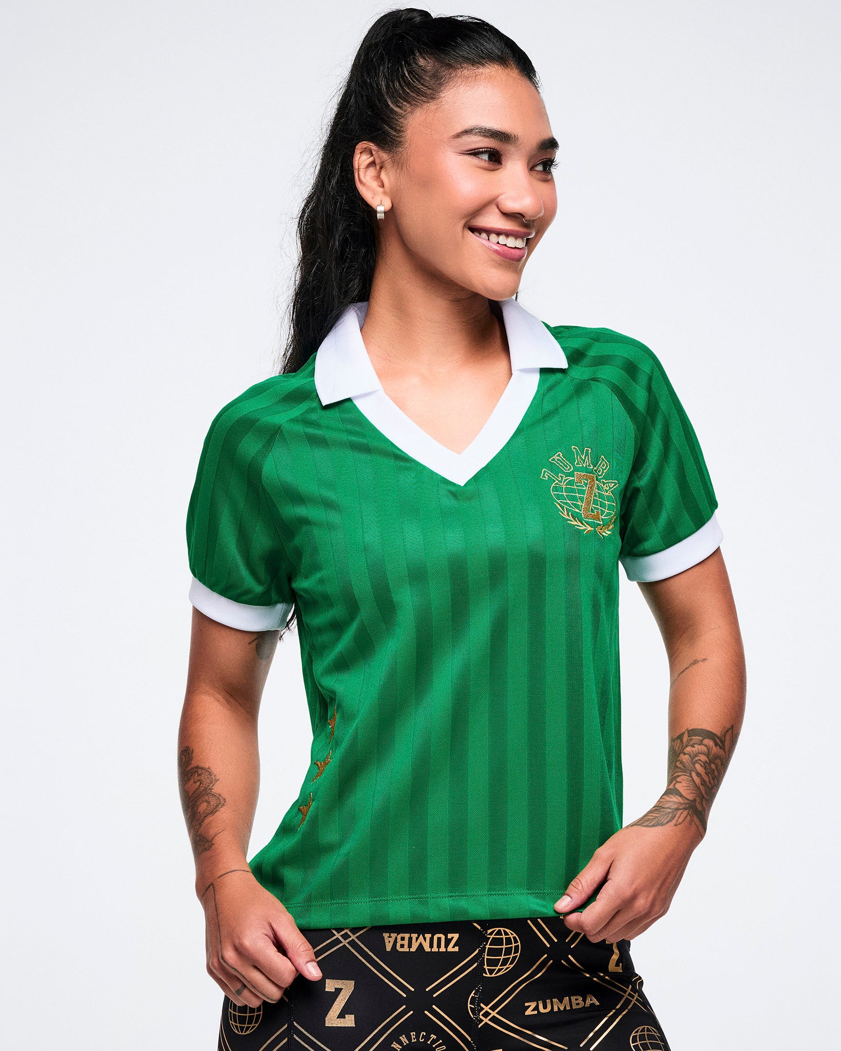 Female model in a green polo with a gold Zumba logo on the top left, white-trim sleeves and collar, on a white background.