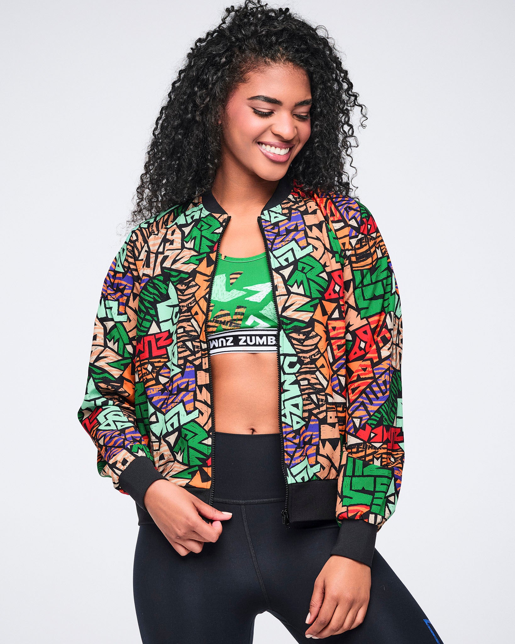 Multicolor Zumba print zip-front jacket with black ribbed trim. Female model wears the jacket open over a green printed sports bra, black high-waisted leggings, and black athletic wear.
