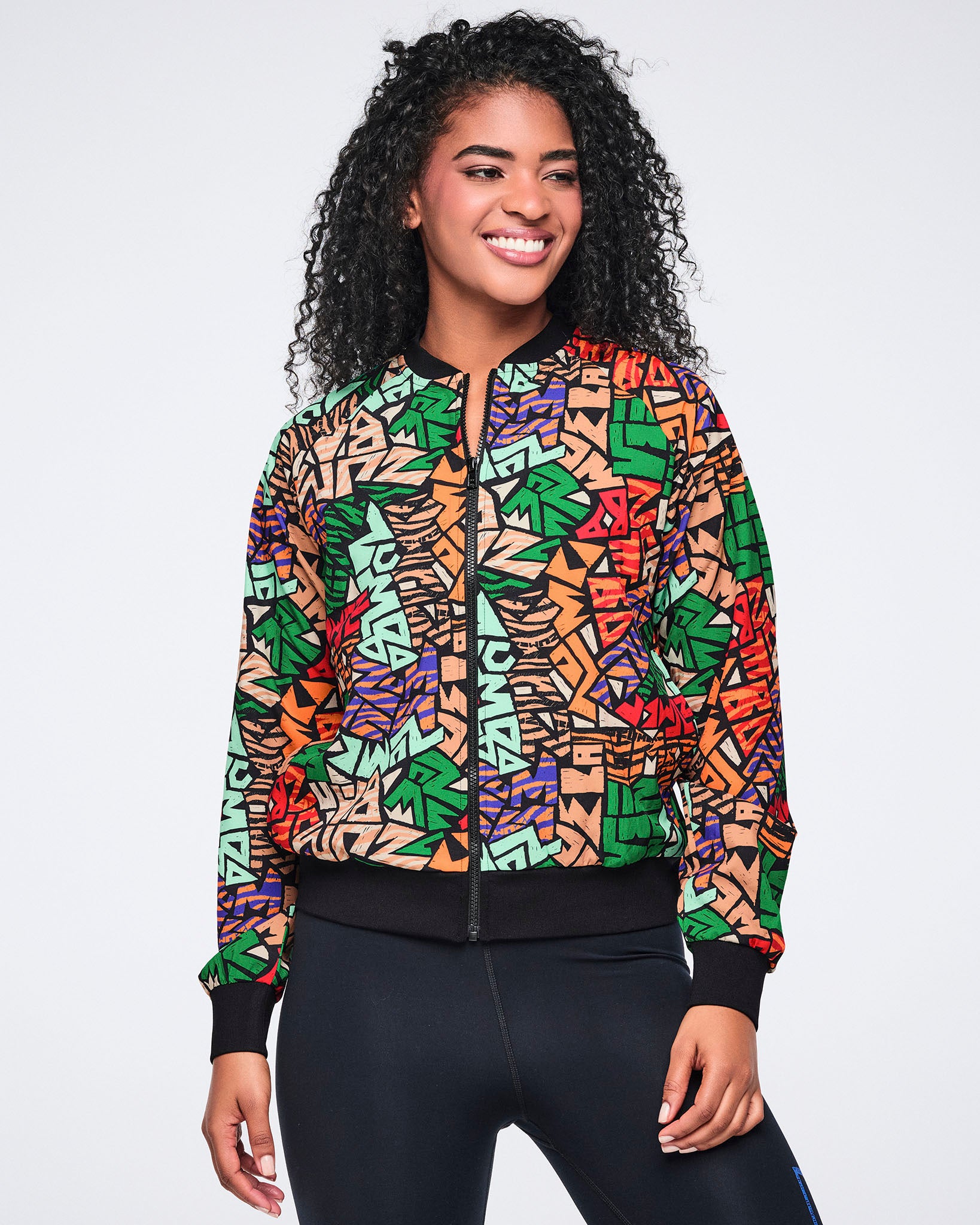 Multicolor Zumba print jacket with a zip front, black ribbed collar, cuffs, and hem. Female model wears the jacket zipped with black high-waisted leggings.