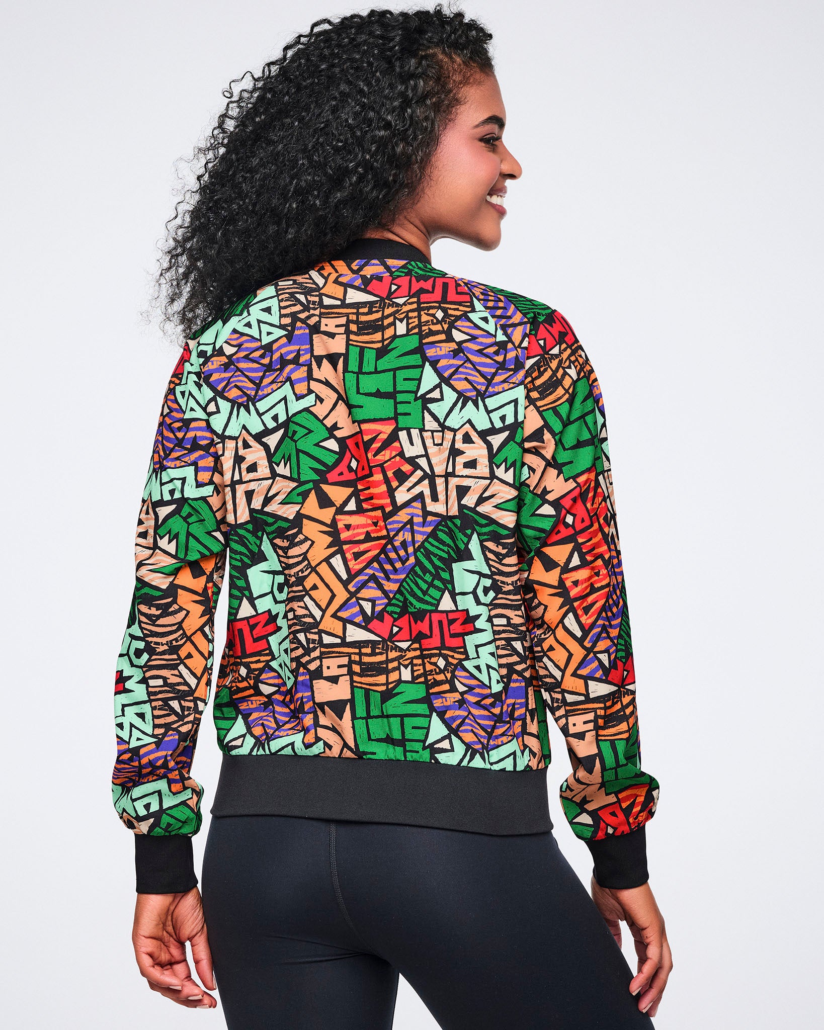 Back view of a multicolor Zumba print jacket with long sleeves, a black ribbed hem, and black cuffs. Female model wears the jacket with black high-waisted leggings, shown from the back.
