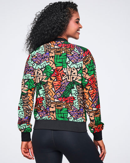 Back view of a multicolor Zumba print jacket with long sleeves, a black ribbed hem, and black cuffs. Female model wears the jacket with black high-waisted leggings, shown from the back.