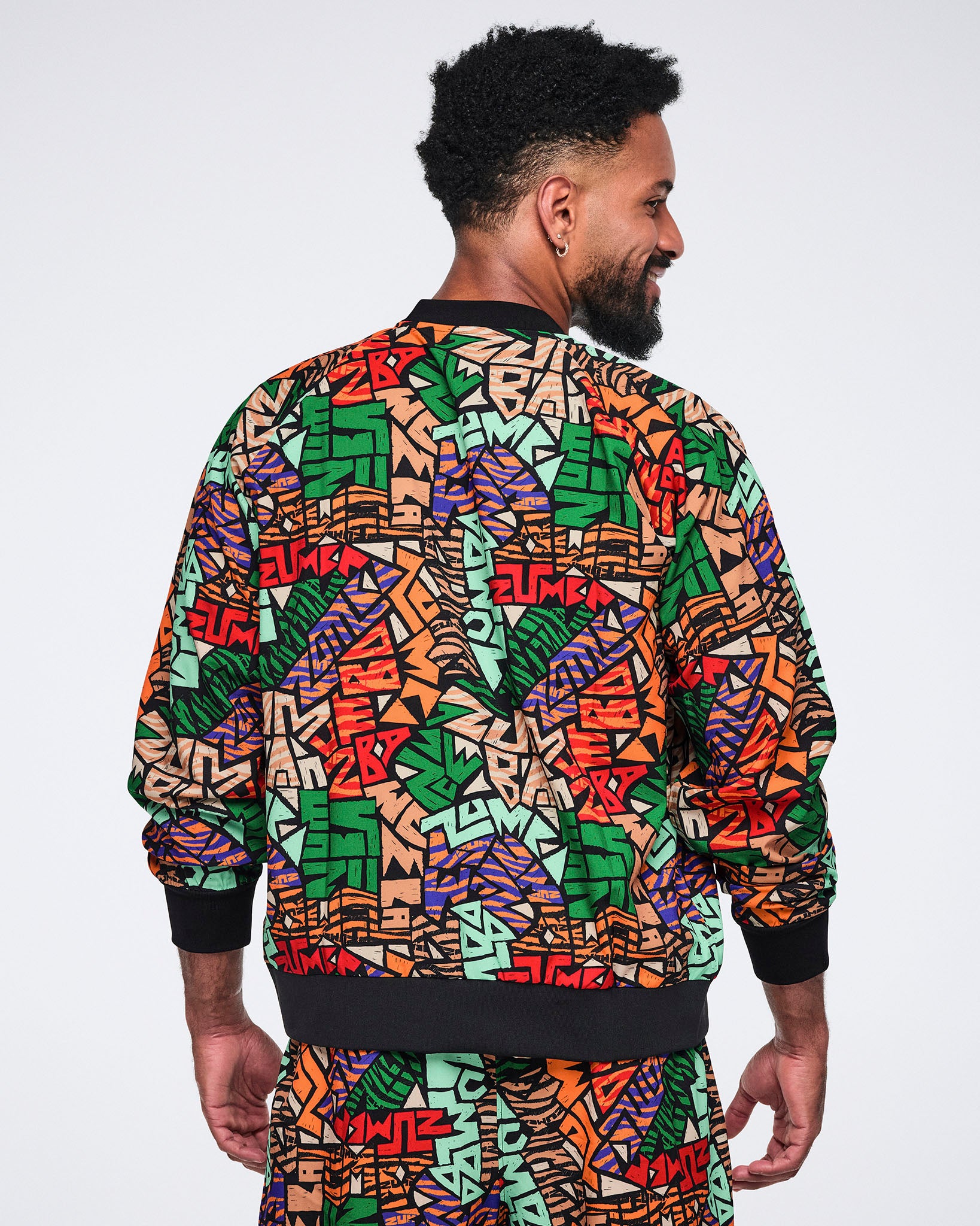 Back view of a multicolor Zumba print jacket with long sleeves and black ribbed hem and cuffs. Male model wears the jacket with matching print pants, shown from the back.