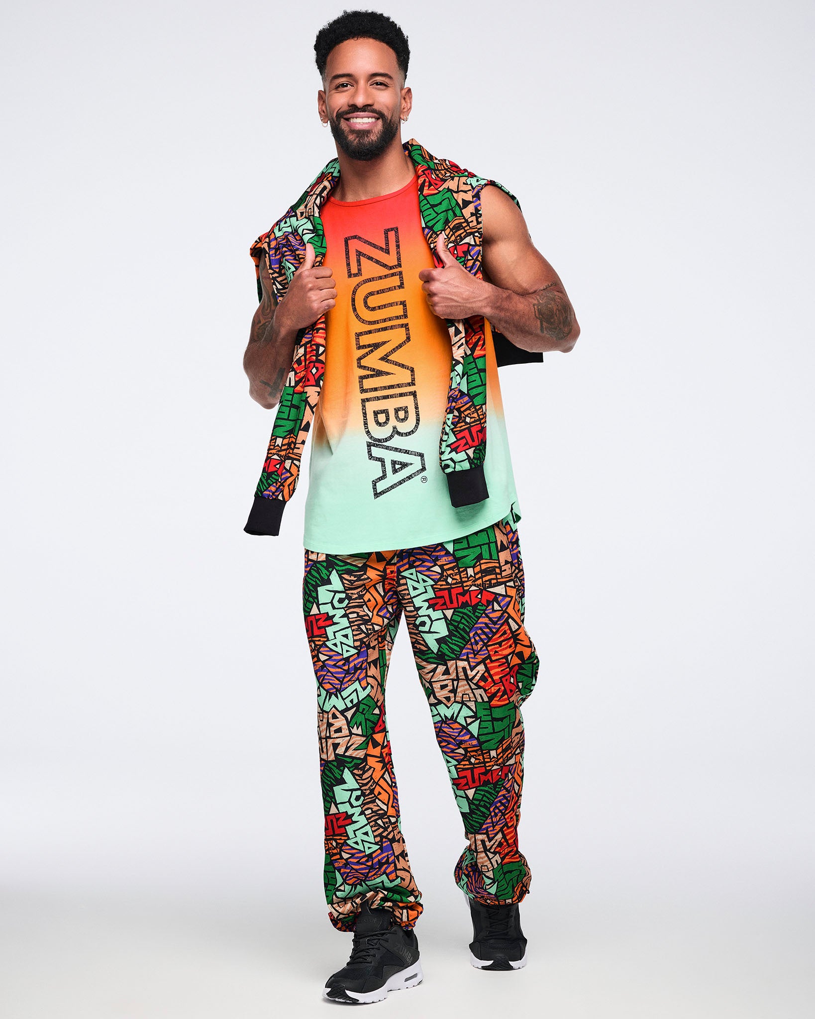 Multicolor Zumba print jogger pants with a relaxed fit and gathered ankles. Male model wears the pants with a sleeveless red, orange and mint gradient Zumba tank, a matching print sweatshirt draped over his shoulders, and black athletic sneakers.