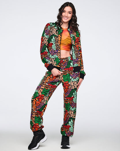 Multicolor Zumba print zip-front jacket and matching jogger pants with black ribbed trim. Female model wears the set with an orange and yellow gradient crop top and black athletic sneakers.