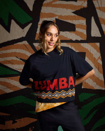 Female model wearing a black oversized cropped T-shirt with red Zumba lettering and an orange, white, and black geometric hem print, styled over a yellow tie-dye layer with black fitted pants in front of a colorful background.