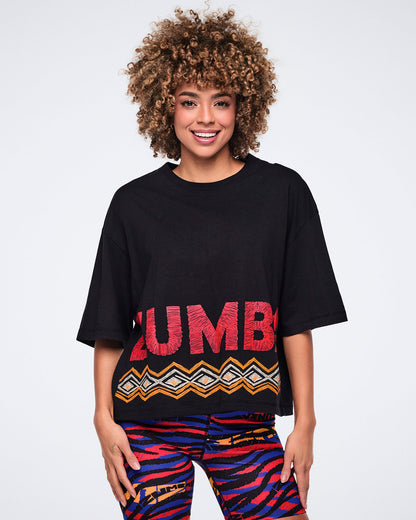 Female model wearing a black oversized cropped T-shirt with large red Zumba lettering and an orange, white, and black geometric border print at the hem, paired with multicolor zebra-print biker shorts.