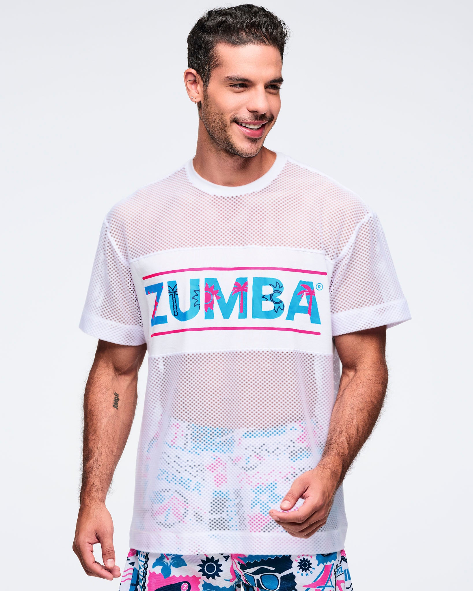 White oversized mesh short-sleeve crewneck top with a bold turquoise “ZUMBA” logo band across the chest and sheer perforated fabric; shown on a male model wearing printed shorts.