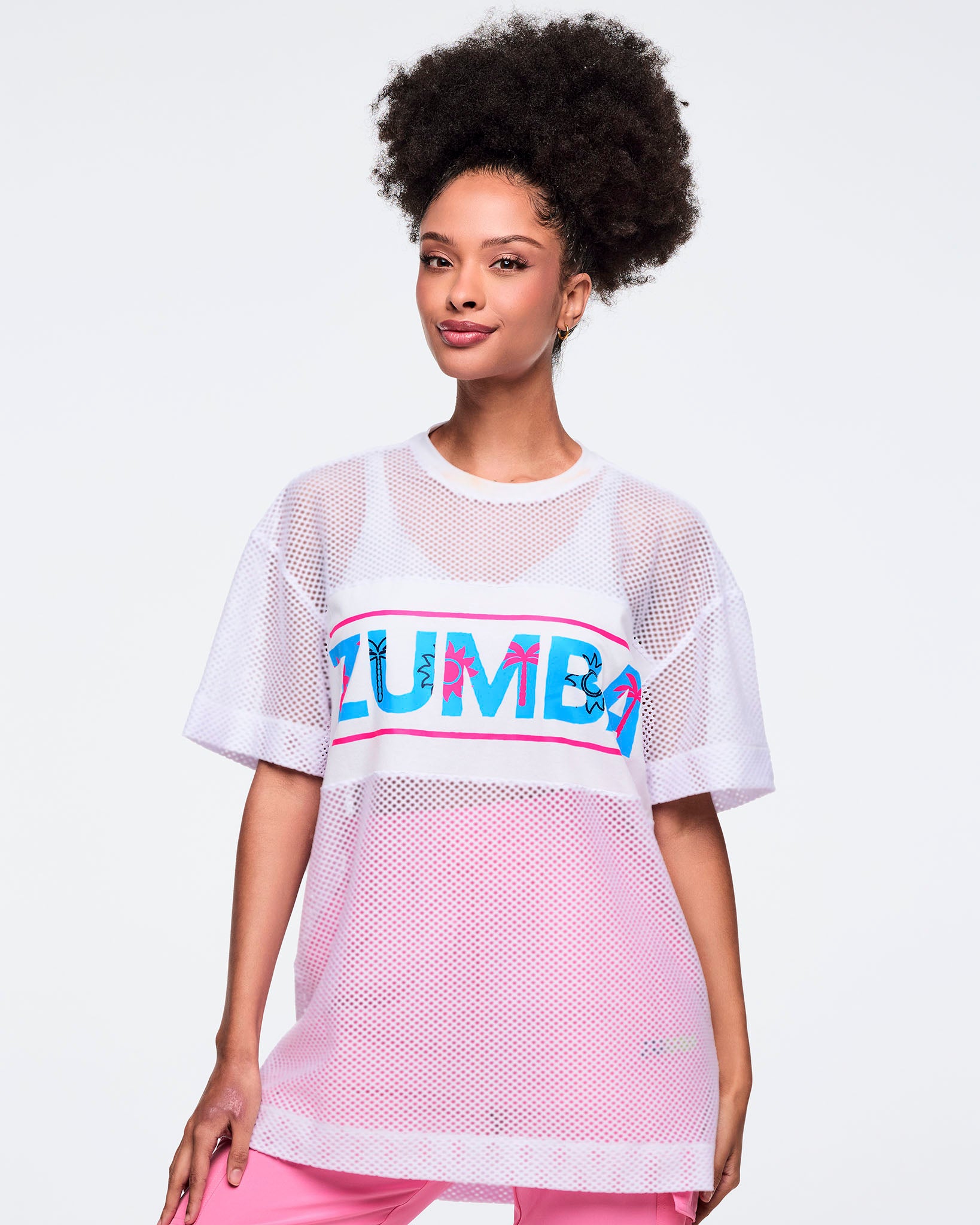 White oversized mesh short-sleeve crewneck top with a turquoise-and-hot-pink “ZUMBA” chest graphic and longline fit; shown on a female model wearing pink pants.