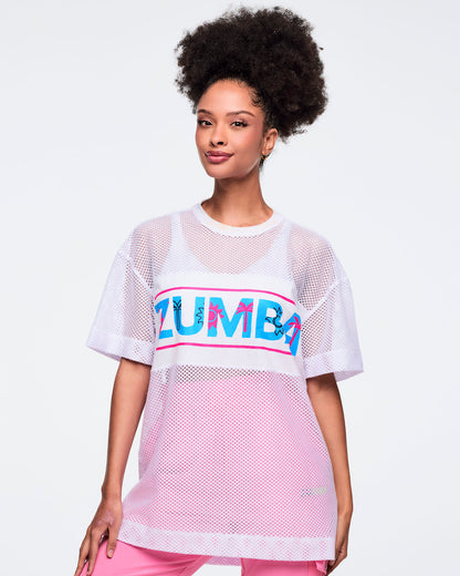 White oversized mesh short-sleeve crewneck top with a turquoise-and-hot-pink “ZUMBA” chest graphic and longline fit; shown on a female model wearing pink pants.