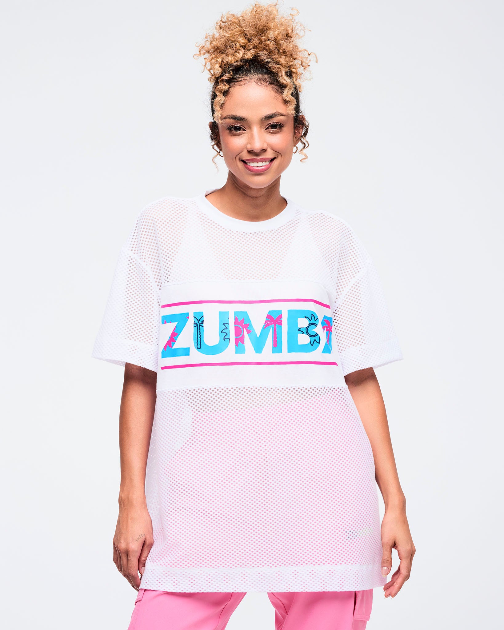 White oversized mesh short-sleeve crewneck top with a wide chest panel featuring a bold “ZUMBA” logo in turquoise with hot-pink accents; shown on a female model wearing pink pants.
